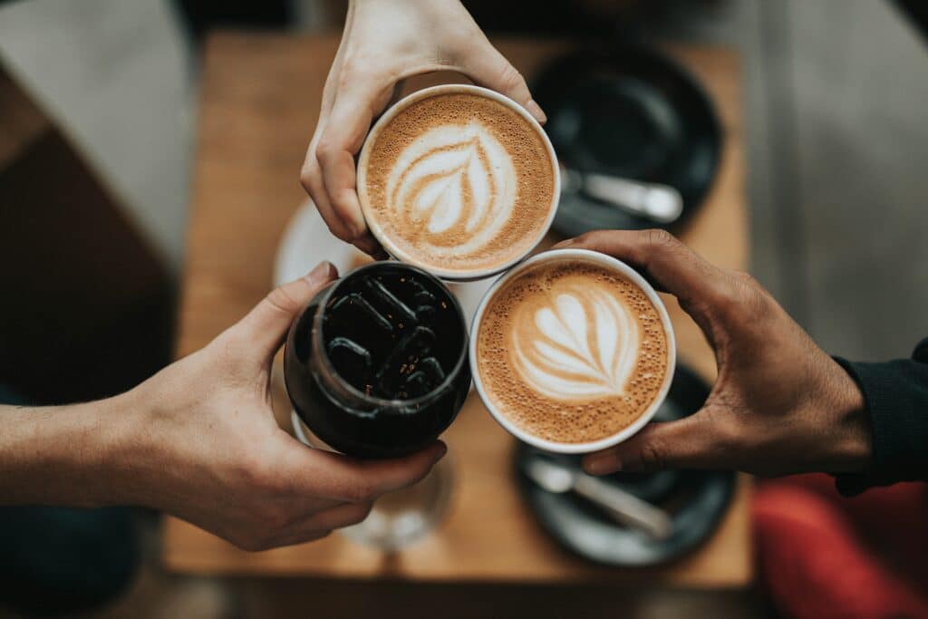 Tre personer skåler med to kopper kaffe med latte art og et glass med en mørk drikk. De sitter ved et trebord, og bildet gir en følelse av samvær og hygge.