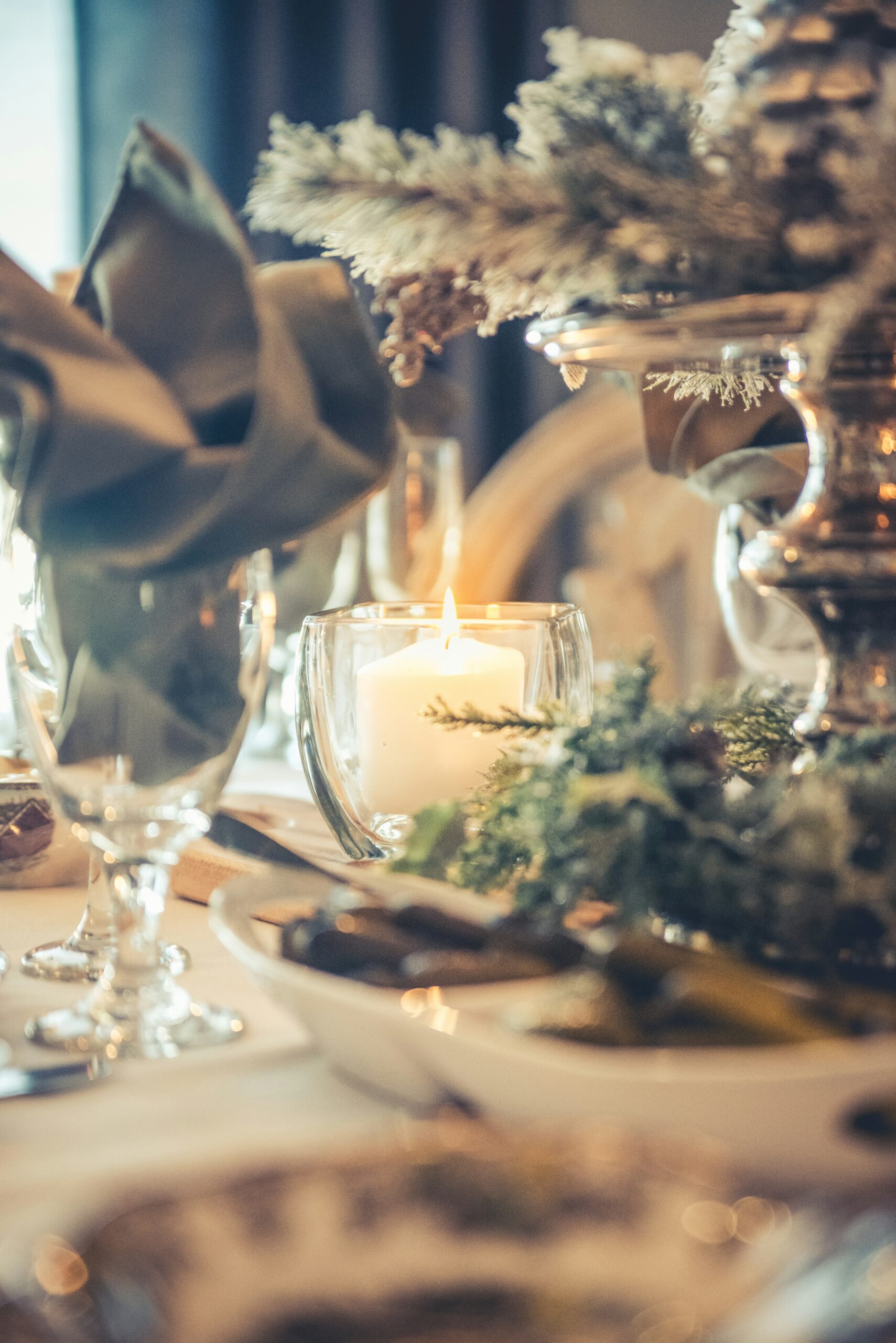 Nærbilde av et elegant dekket julebord med et tent stearinlys i en glasslysholder i sentrum. Bordet er pyntet med granbar og dekorasjoner, og glassene er pent plassert rundt. Stemningen er varm og innbydende, med en koselig og festlig atmosfære.