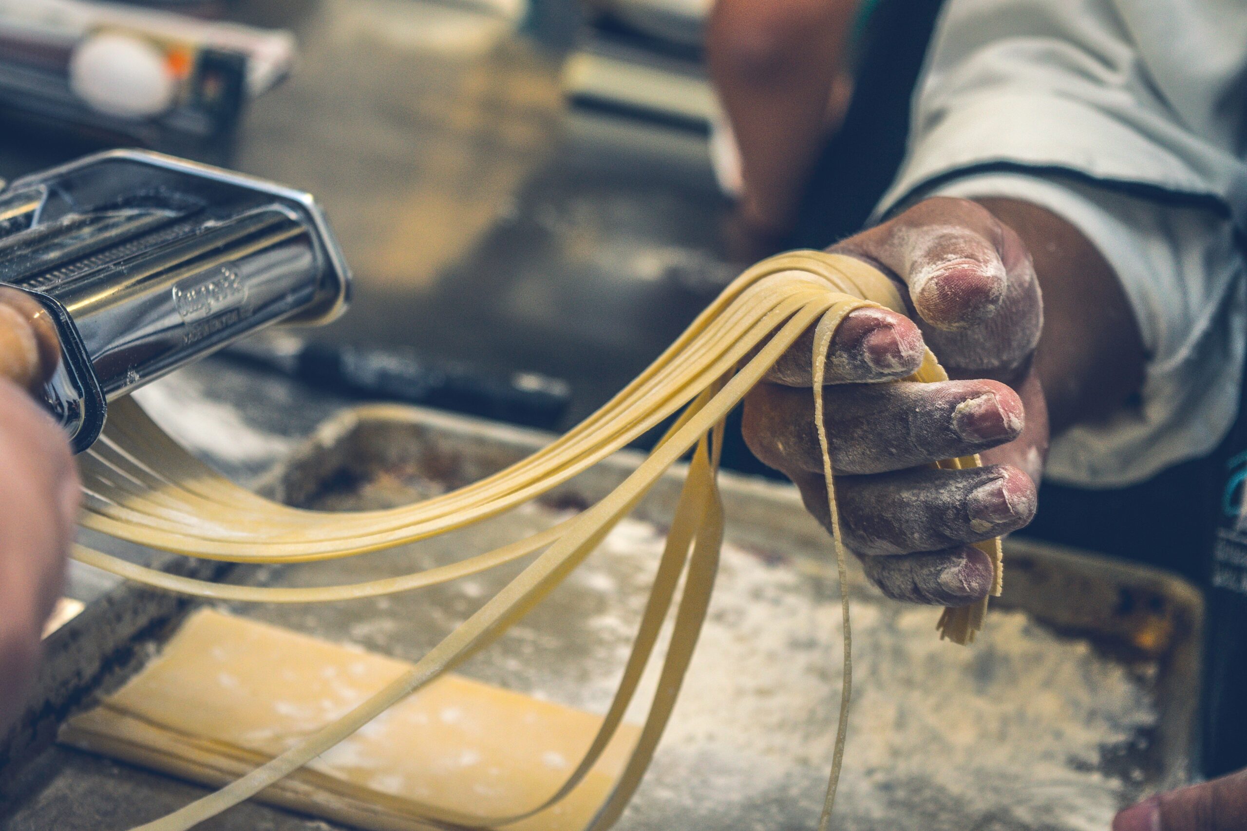 Nærbilde av en person som lager fersk pasta. Hendene, dekket med mel, trekker lange, tynne pastastrimler gjennom en pastamaskin. Det er mel på arbeidsflaten, og pastadeigen ser fersk og hjemmelaget ut.