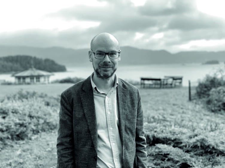 Svart/hvitt foto av Øivind Torkelsen, forsker på stamcellebehandling og nevrolog. Han står på en strand med sjøen i bakgrunn.